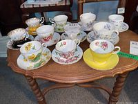 Full view of 11 assorted fine bone china teacups and matching saucers arranged on wooden table featuring floral patterns, various colors including yellow, pink, blue, and white.