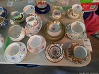 Overview of the 9 teacups with saucers, creamers, sugar bowls, and a plate showing variety of brands and designs