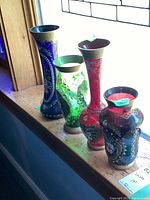 Four ornate colored glass vases lined up on a window sill, showing different sizes and hand-painted floral designs with gold accents.