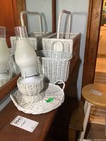 Photo showing all four items: white wicker magazine rack, basket, round tray, and oil lamp with wicker base and glass chimney.
