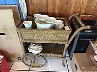 Front angle photo of the vintage wicker cart showing the basket top, lower shelf, and one large spoked wheel. Partial view of surrounding room and boxes.