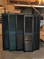 Five antique wooden shutters standing upright in a basement. Various dark colors, mainly black and green, showing signs of wear and age.