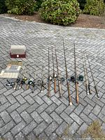 Overview of all fishing rods with reels arranged on pavement alongside tackle and fly boxes. Shows quantity, variety, and condition of rods and reels.