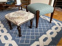 Two footstools side by side on blue and white patterned rug. Smaller stool has floral upholstery and turned dark wood legs. Larger stool has blue upholstery with geometric pattern and straight wooden legs.
