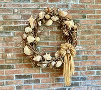 Front view of large grapevine wreath with multiple seashell varieties and a straw bow, hanging on brick wall