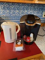 Keurig coffee maker and electric tea kettle on a red countertop with blue patterned wallpaper background.