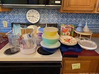 Wide view of the large assortment of kitchen plastic serving and storage items arranged on stovetop and counter including bowls, plates, organizers, trays, funnels, and lazy susan.