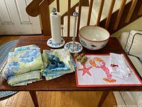 Photo of hydrangea print tablecloths and napkins, lobster themed placemat and white linen napkin, brass shell napkin rings, ceramic paper towel holders, and large ceramic bowl on wooden table