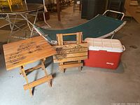 Photo showing green fabric hammock on metal stand, two wooden folding tables with faded ruffed grouse designs, red Gott cooler, small waste basket on concrete floor
