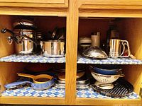 Overview showing multiple stainless steel pots and pans, enamel colanders, and various kitchen tools stored in wooden cabinet shelving.
