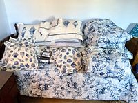 Wide view of bedding lot with folded comforters, pillow shams, throw pillows, chair pads, and other textile items arranged on a wooden bench.
