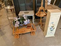Various small tables, trays, cabinet, folding lap tray, cranberry rake, faux flower display and more, shown against a backdrop in a basement location. Items show wear and need cleaning.