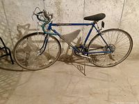 Side view of vintage blue Schwinn Varsity 10 speed bike leaning against concrete wall in basement, showing overall frame, wheels, seat, and drivetrain