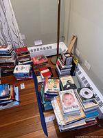 Multiple stacks of books on the floor near a lamp and corner wall, containing children's books, notebooks, and other reading materials.