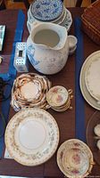 Photo showing stacks of plates with blue floral patterns, Continental Ivory cups and saucers with floral and gold details, and the white and blue porcelain pitcher with visible damage.