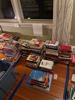 Wide shot showing many stacks of books on a wooden floor near windows.