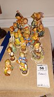 Full view of all nineteen Hummel figurines arranged on a beige cloth, showing variety of figures and characters in the collection.