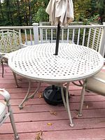 Photo showing round white metal outdoor table with weathered lattice top and attached umbrella with beige cover. Matching metal chairs with beige cushions placed around table on wood deck.