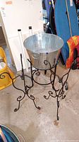 Front view of two black metal plant stands and large aluminum bucket on floor.