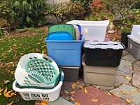 Photo of various storage bins stacked outdoors on stone surface including blue, beige, black, gray, and purple colors with some bins covered with lids and some open. Two white laundry baskets and a green laundry basket insert shown nearby.