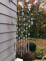 Two wind chimes hanging outside on a wall; one with silver tubes, green beads, and wooden balls, and the other with bronze tubes on a wooden ring.