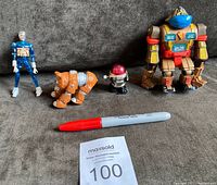 Four vintage action figures arranged on a gray surface with a red marker for scale: a blue humanoid figure, orange Daggit, red-headed small robot, and large beige and red robot.