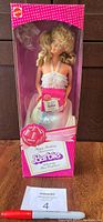 Front view of unopened pink polka dot box showing Barbie doll with blonde curly hair and white halter dress inside, holding a small pink box.