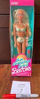 Front view of 1991 Sun Sensational Barbie Doll in original unopened box with pink top and green background inside the box. Doll has real lipstick and wears gold bikini with necklace and earrings visible.