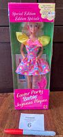 Front view photo of the Barbie doll in original packaging showing the doll in a colorful floral dress with yellow puffed sleeves, pink bow, and visible accessories including egg holder and dye.