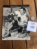 Full view of black and white photo showing Glenn Hall in goalie gear with autograph in gold sharpie and quarter placed beside for scale