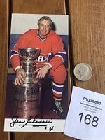 Photo of Jean Beliveau in Canadiens uniform posed with Stanley Cup next to a coin for scale and auction tag.