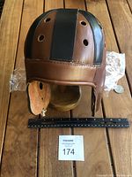 Front view of brown and black leather reproduction vintage football helmet on wooden stand with ruler for scale