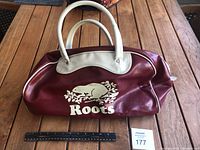 Overall front view of the red leather Roots carry bag showing the white beaver logo and cream handles. Measures roughly 18 inches in length.