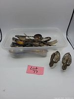 Plastic container holding various antique forks and spoons, showing tarnished metal condition, along with two metal salt cellars with swan motifs next to the container.