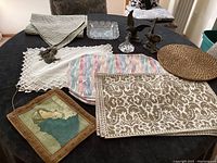 Overview of all items on a black table including brass eagle bookends, various fabric linens, cut glass paperweights, and woven tray.