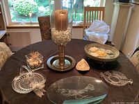 Overview of all items on black table including large glass candle holder, oval etched glass tray, two glass candy dishes, ceramic bowl on stand, glass cube container, and rock.