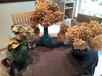 Photo showing three glazed ceramic vases in blue and green tones and two clear glass smaller vases, all holding dried hydrangea flower bunches