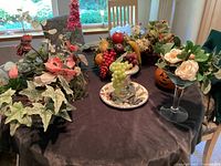 Wide view of several kitschy faux flower and fruit arrangements on a dark tablecloth including Halloween gourd and glass vessel with flowers.
