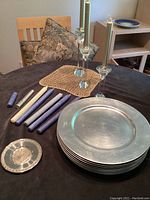 Photo showing 8 large silver lacquer plates stacked and grouped with purple and gold taper candles alongside two glass candle holders on a dark table