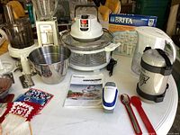 Photo of Sunbeam food processor with stainless bowl, blender attachment, Decosonic convection roaster oven, Brita pitcher and unopened filters, kitchen scale, and various kitchen utensils on a white table.