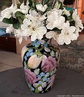 Front view of ceramic vase with painted floral design and white artificial flower arrangement on top in indoor setting.