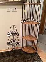 Two corner display racks and a plant stand shown together against wall indoors on carpet. Tall rack with wicker shelves and grapevine metal decorations on top and sides, shorter completely metal corner rack with curled decorative pattern, and small black metal scrollwork plant stand.