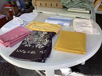 Overview of 7 folded tablecloths on white round table, showing mix of vintage floral, lace, and solid colors including yellow, pink, white, and blue.