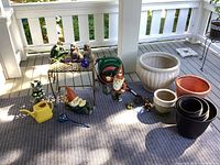 Overall view of assorted yard decor and garden accessories laid out on porch including gnomes, frogs, pots, watering can, hose and table.