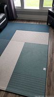 Photo showing full view of area rug with green, beige, and blue blocks and striped detail near window and furniture