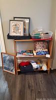 Photo of small shelving unit holding yarn balls in multiple colors, knitting magazines, plastic containers with craft supplies, and two framed needlepoint artworks stacked in a black basket on top.
