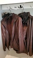 Two brown Danier leather jackets hanging side by side with three pairs of black leather gloves on the hangers.