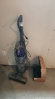 Full view of upright Dyson vacuum cleaner with cord wrapped around handle and cardboard box of attachments on floor
