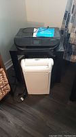 Black HP Envy 4516 printer on top of a small black table with white paper shredder below the table