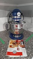 Front view of blue stand mixer on countertop with bowl and pouring shield attached and manual in front